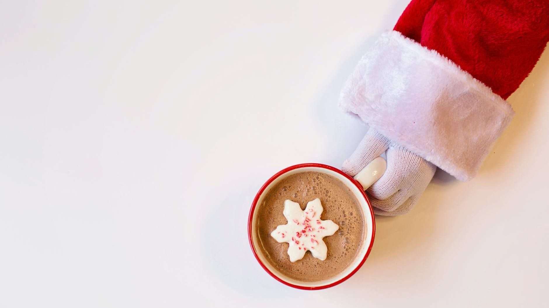 mug of chocolate drink with snowflake shaped cookie on top held by a person in santa suit