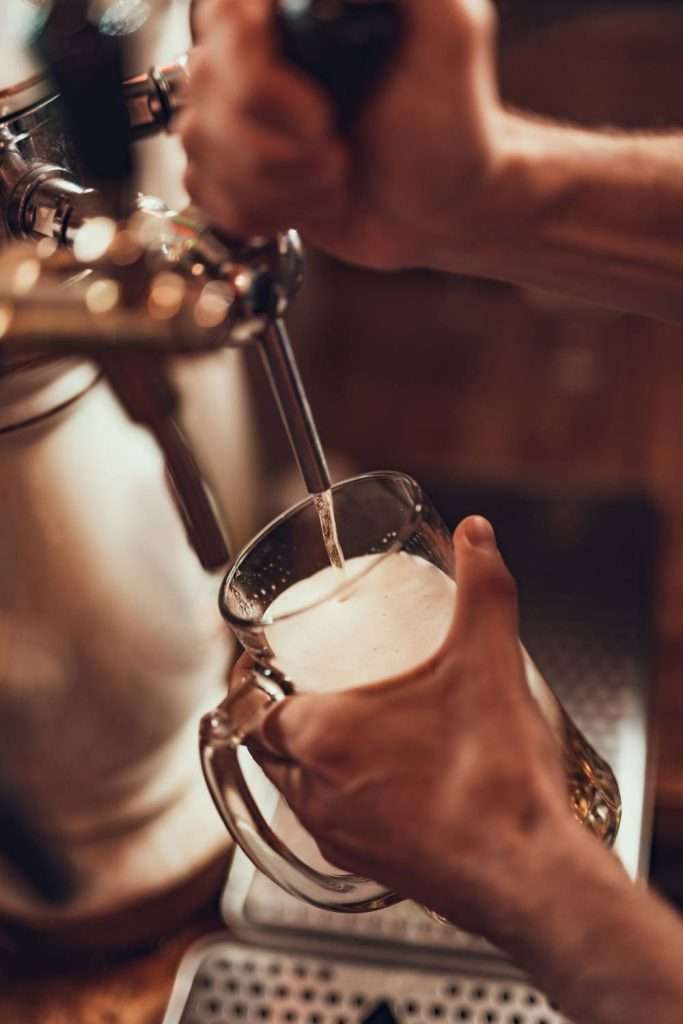 Close-up of a bartender's hands pouring a golden and bubbly draft beer into a glass over a wooden counter.