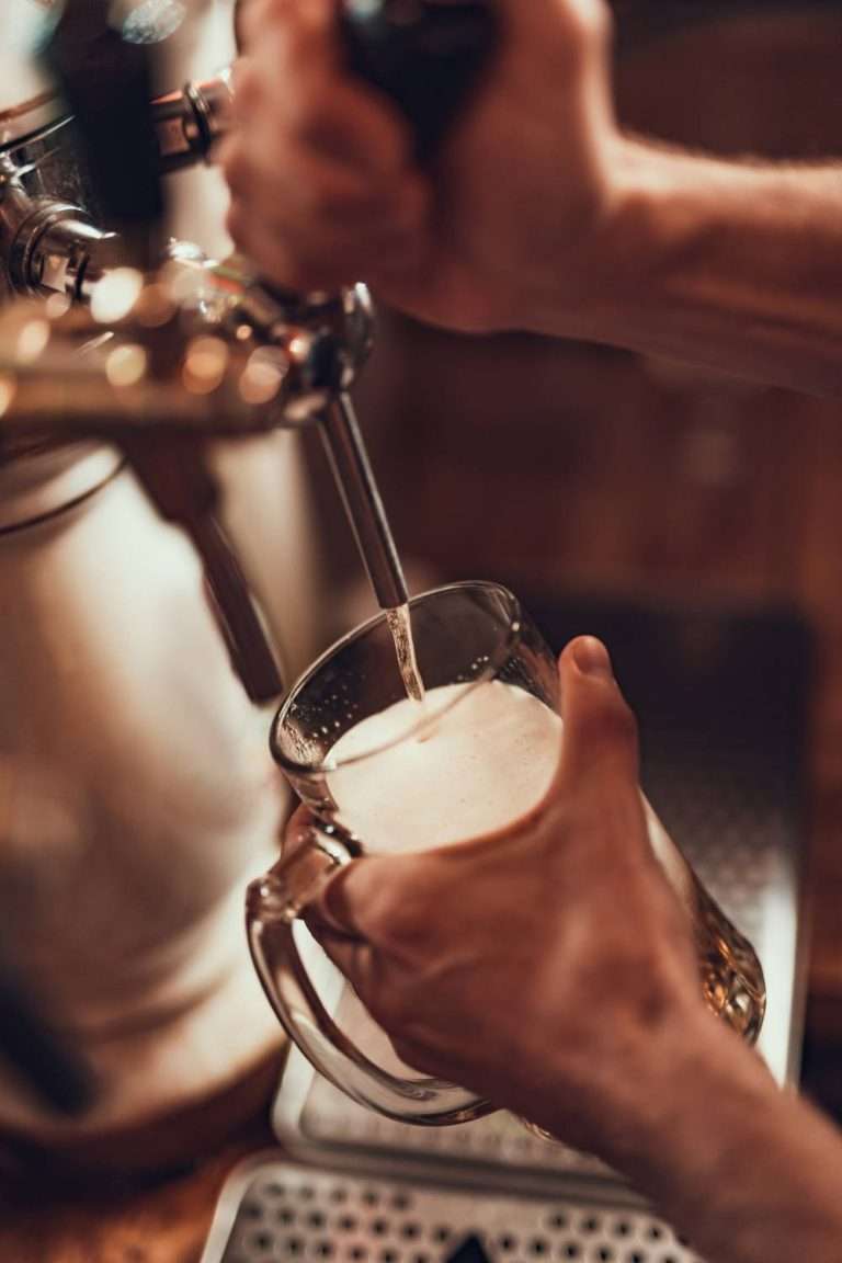 Close-up of a bartender's hands pouring a golden and bubbly draft beer into a glass over a wooden counter.