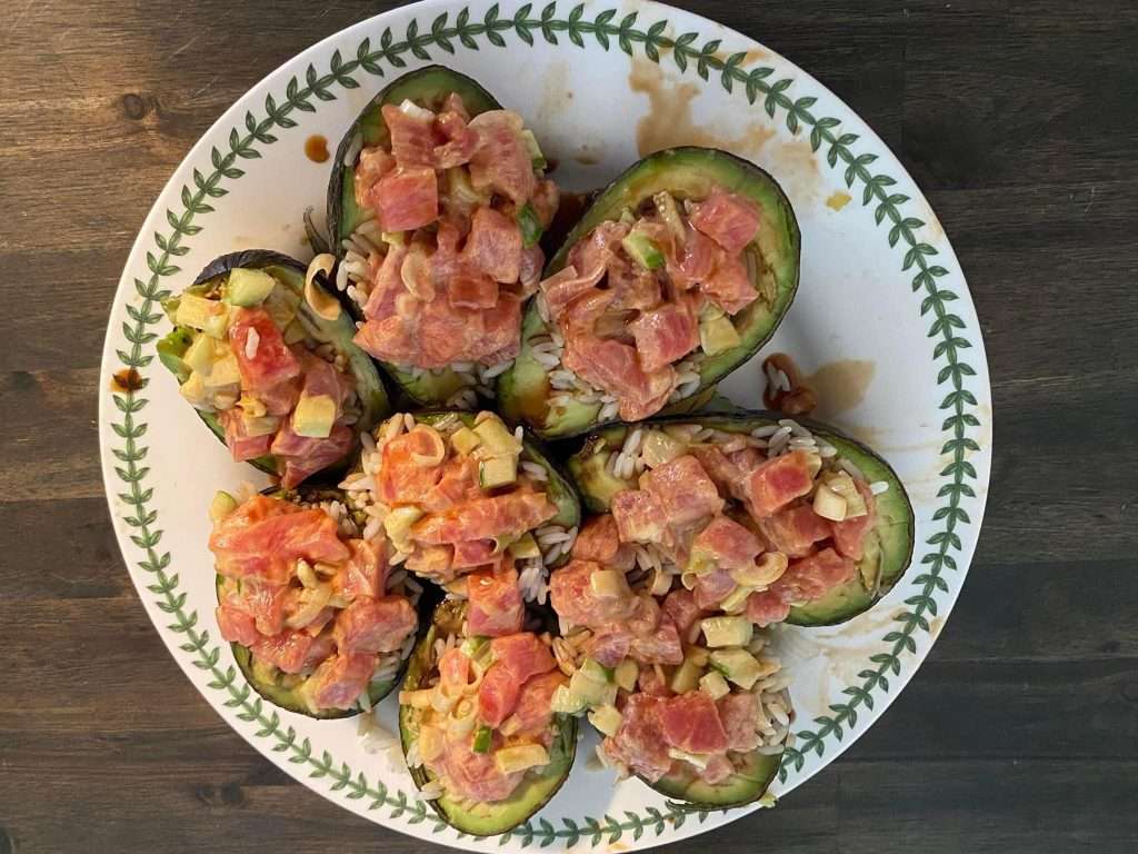 A plate of stuffed avocado with the interior filled with tuna and rice placed in a white dish that has green lines around it, it's sitting on a wood table, the shot was taken from above at eye level, it looks delicious and very appetizing, the colors are vibrant and saturated, the photo is in the style of hyper realistic photography. 