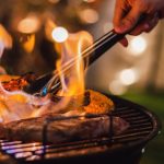 A close-up of someone's hand cooking barbecue and grilled meats on an outdoor camping grill at night time.