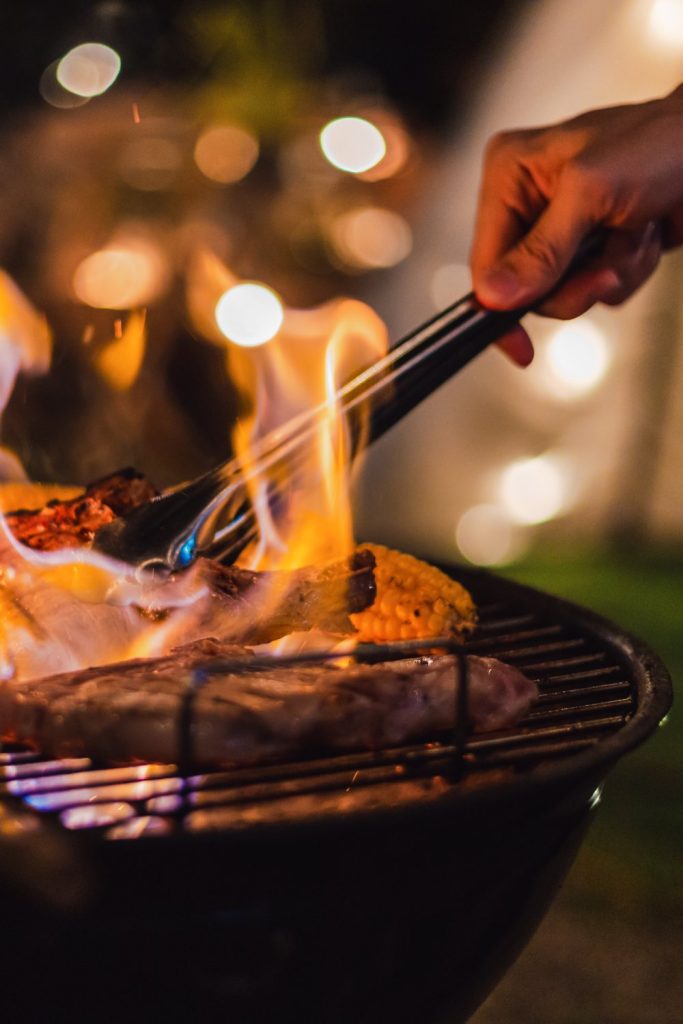 A close-up of someone's hand cooking barbecue and grilled meats on an outdoor camping grill at night time.