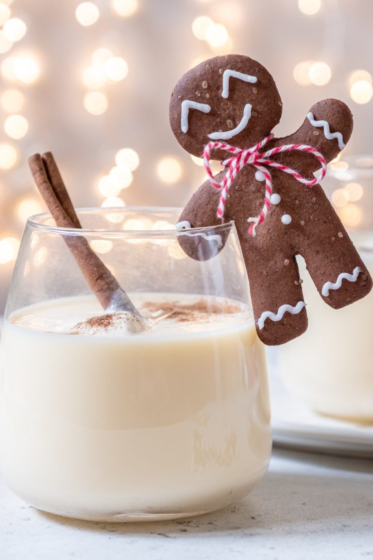 Two glass cups filled with a white liquid and a cinnamon stick sit on a white table. White fairy lights are behind the cups.