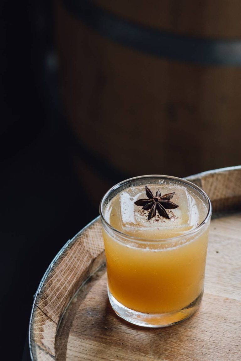 A small cocktail glass filled with a golden-colored drink, ice cube, and star anise garnish on a wooden barrel.