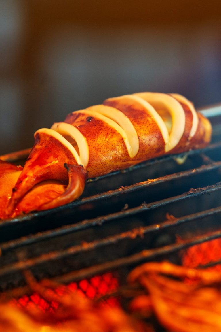 A close-up or a whole squid head with horizontal slices through the flesh sitting on a hot grill alongside other meat pieces.