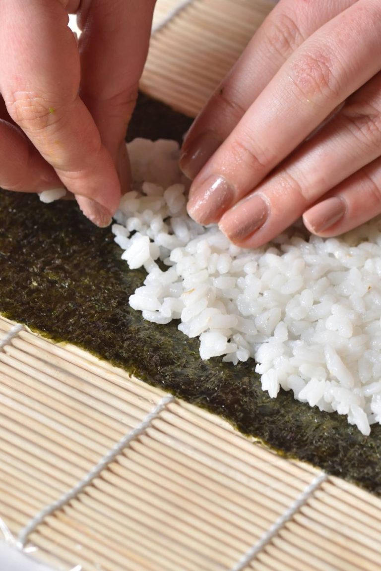 A person is using their hands to press white rice onto a piece of seaweed that's on top of a plastic-wrapped sushi roller.