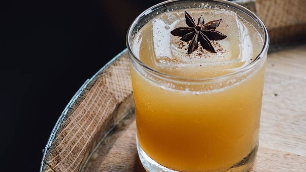 A small cocktail glass filled with a golden-colored drink, ice cube, and star anise garnish on a wooden barrel.