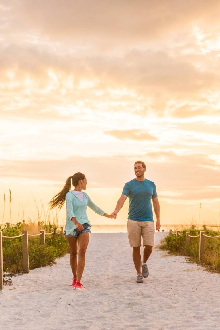 A happy couple hold hands while walking up a sandy path away from the ocean at sunset. A fence and greenery line the path.