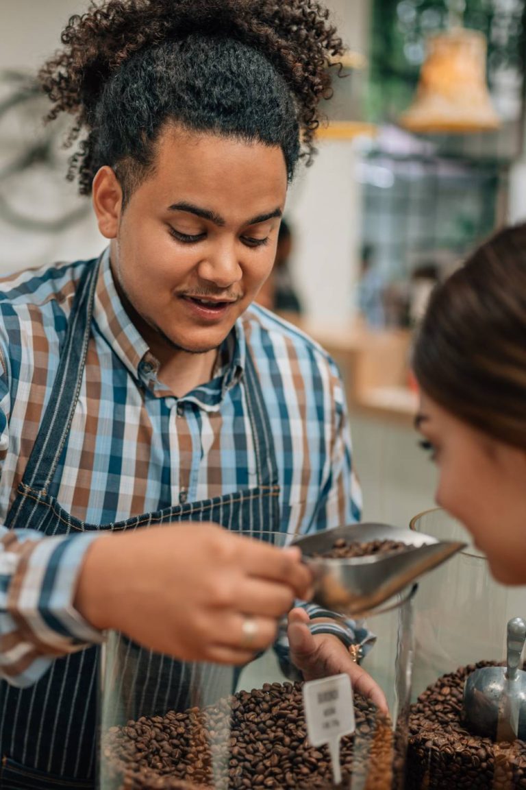 A coffee shop worker in an apron lifting a scoop of roasted coffee beans to allow a customer to smell the roast.