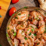 Pieces of raw, marinated chicken in a glass bowl, sitting next to a wooden cutting board, a knife, and spare ingredients.