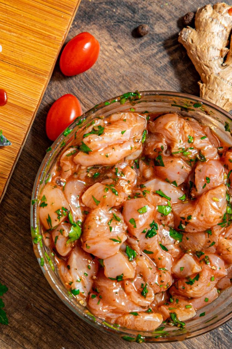 Pieces of raw, marinated chicken in a glass bowl, sitting next to a wooden cutting board, a knife, and spare ingredients.