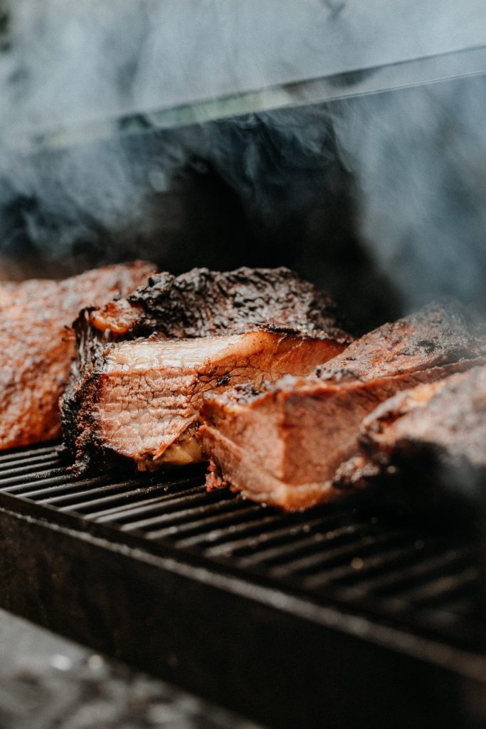 A close-up view shows five large pieces of meat sitting on a black smoker with smoke rising from the meat.