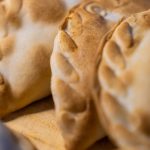 A close-up of four empanadas under dramatic lighting, served on a wooden board. The pastry looks crispy and brown.