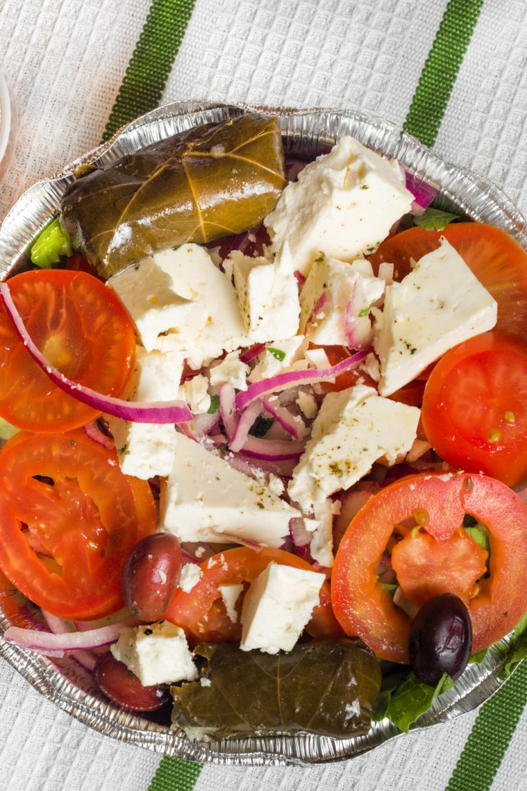 A Greek salad, consisting of lettuce, tomatoes, and feta cheese, sits in an aluminum container on a table next to a sauce.