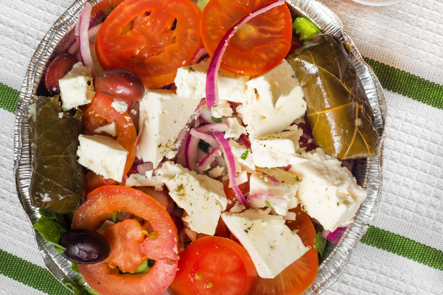 A Greek salad, consisting of lettuce, tomatoes, and feta cheese, sits in an aluminum container on a table next to a sauce.