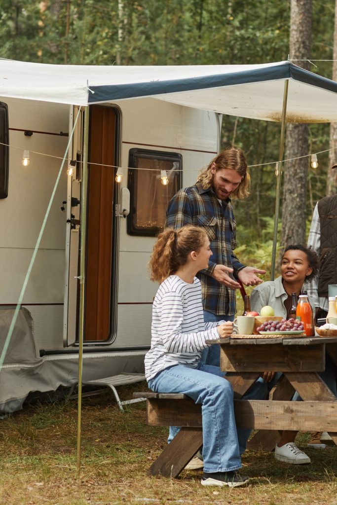 A group of five people gather around a wooden picnic table next to a parked camper van with an awning.