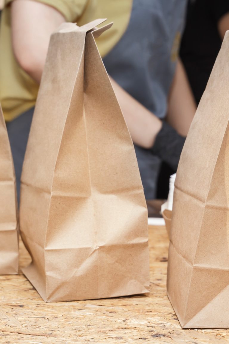 Three folded brown paper bags sit on a counter in front of people, with cardboard containers partially in view.