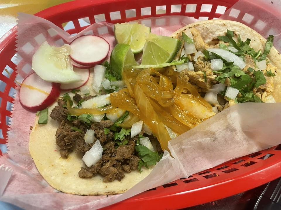 Close-up of steak and chicken street tacos topped with onions and cilantro, served with grilled onions, radishes, cucumber slices, and lime wedges.
