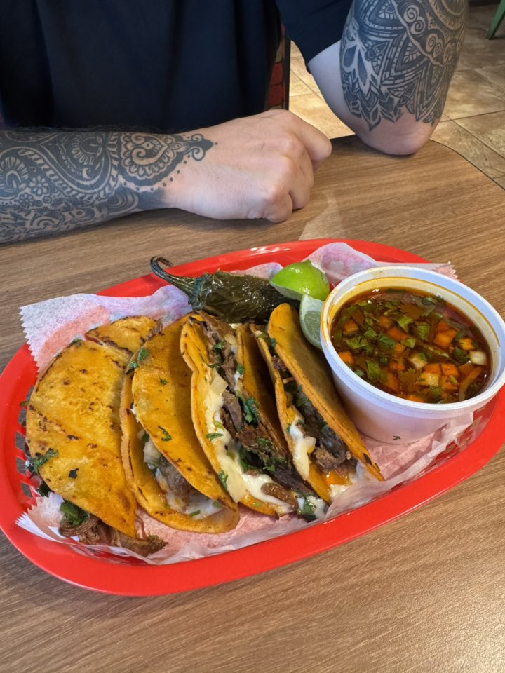 Four crispy birria tacos with melted cheese and beef served with lime wedges, grilled jalapeño, and a side of consommé.