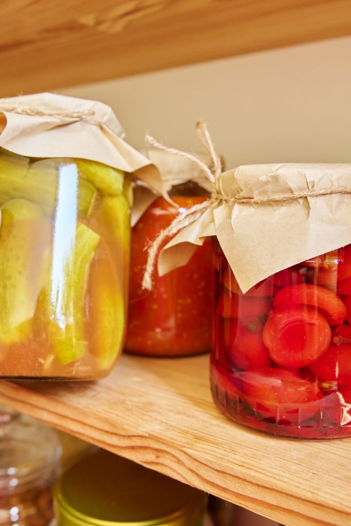 A hand is grabbing a large jar of preserved pickles from a pantry shelf full of additional preserved foods.