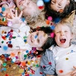 Four cheerful children lying on the floor, playfully surrounded by vibrant confetti.