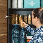 A woman with a ponytail stands in front of an open cabinet with several containers of dried foods, bottled water, and supplies.