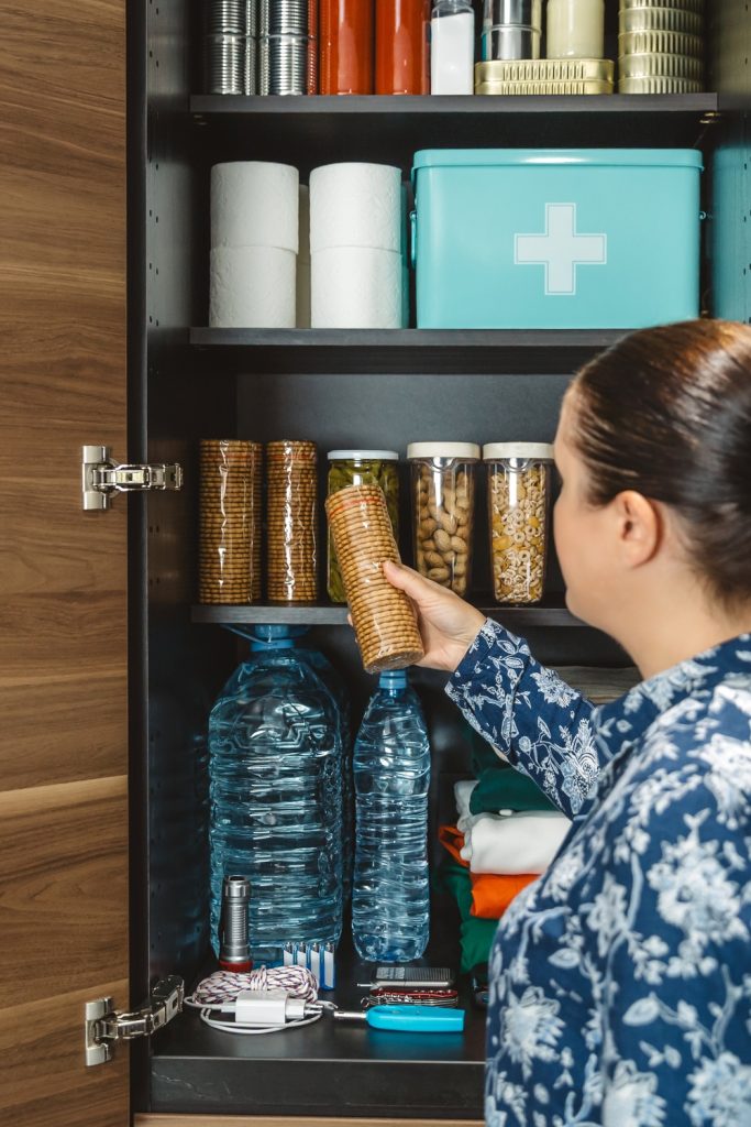 A woman with a ponytail stands in front of an open cabinet with several containers of dried foods, bottled water, and supplies.