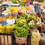A fully stocked fruit and vegetable stand at a farmer's market includes piles of bananas under a rainbow umbrella.