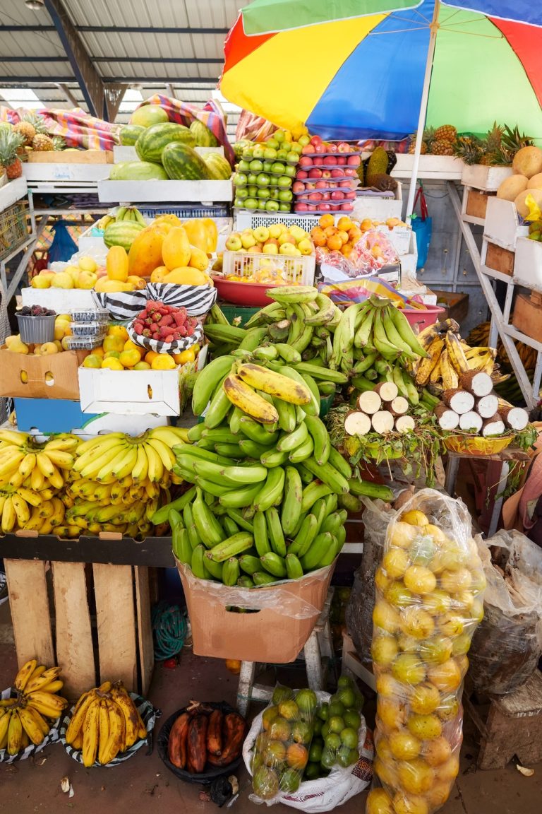 A fully stocked fruit and vegetable stand at a farmer's market includes piles of bananas under a rainbow umbrella.
