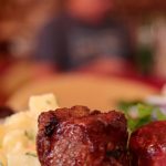 A glass of wine is on a table next to a plate filled with mashed potatoes, steak, and greens. A fork is in front of the plate.