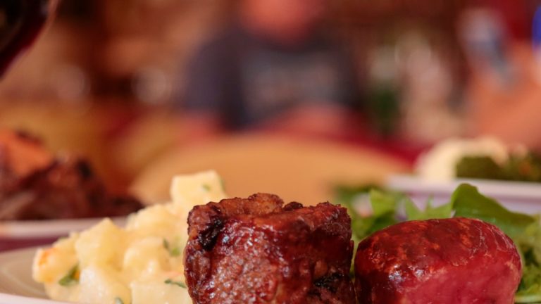 A glass of wine is on a table next to a plate filled with mashed potatoes, steak, and greens. A fork is in front of the plate.